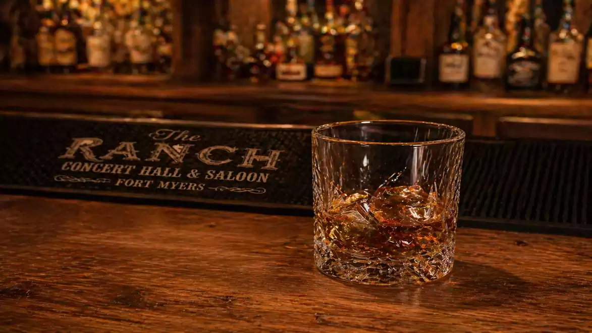 A glass of whiskey with ice cubes sits on a wooden table in a bar, with bottles and a sign reading 'The Ranch Concert Hall & Saloon' visible in the background.