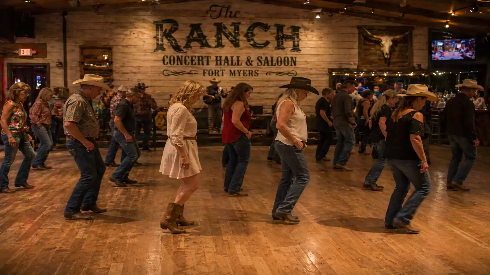 Group of people line dancing in a country bar with wooden floor and cowboy hats.