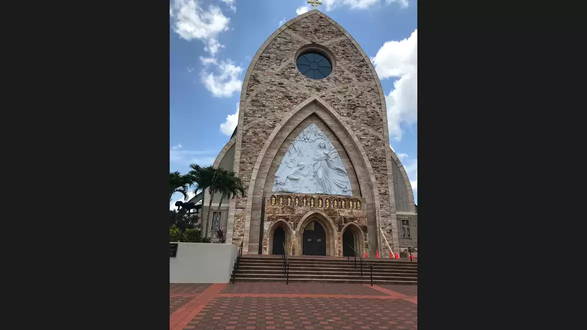 Front view of a stone church with large, intricately designed wooden doors and a relief above the entrances. The sky is clear with a few clouds.