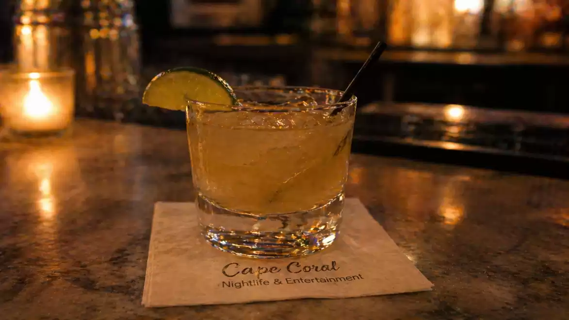 A cocktail glass with a drink, garnished with a lime slice, sits on a marble counter in a dimly lit bar.