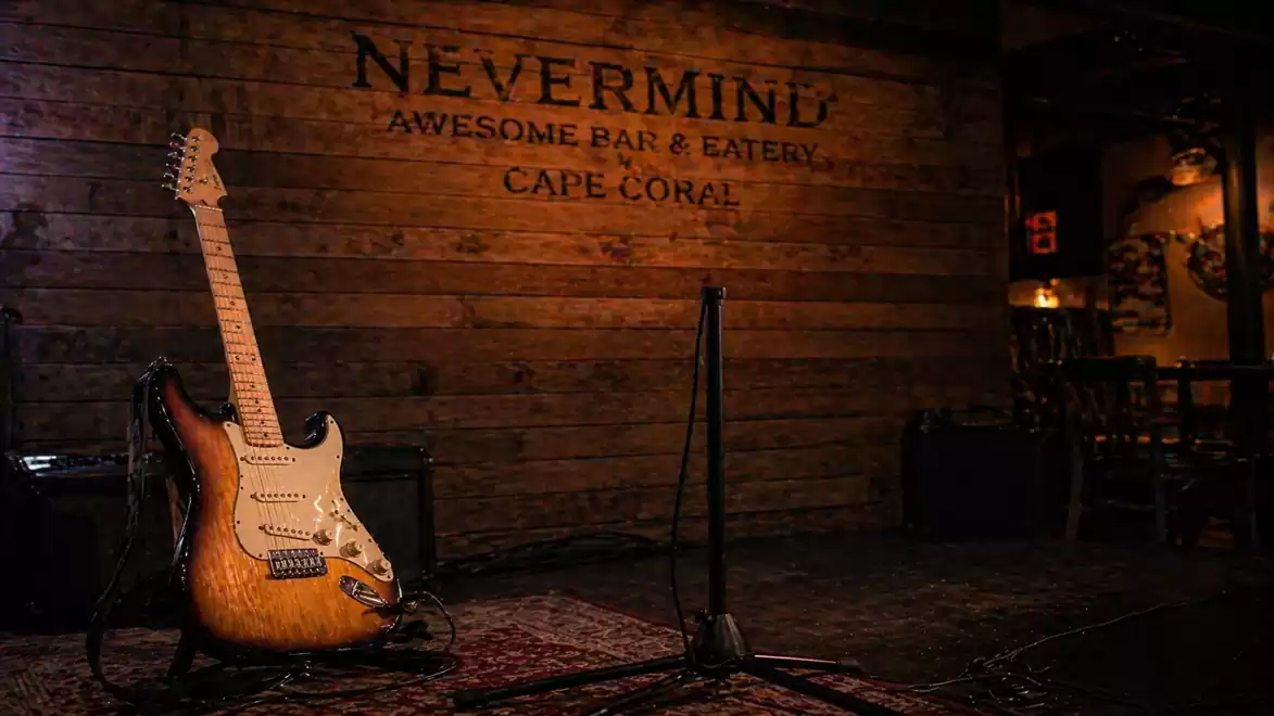 An electric guitar leans against a microphone stand on a stage, with a wooden background displaying 'Nevermind Awesome Bar & Eatery Cape Coral'.