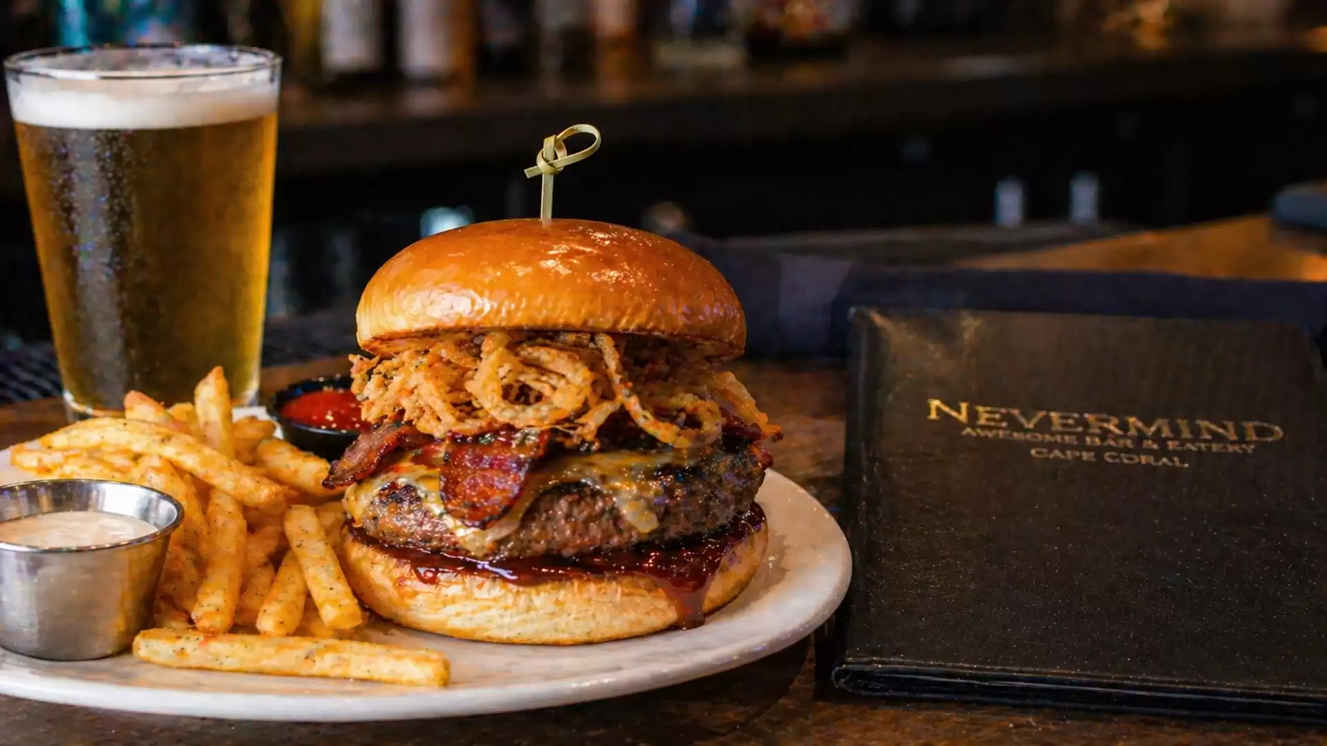 Juicy burger with onion rings, bacon, and fries on a plate next to a glass of beer in a restaurant.