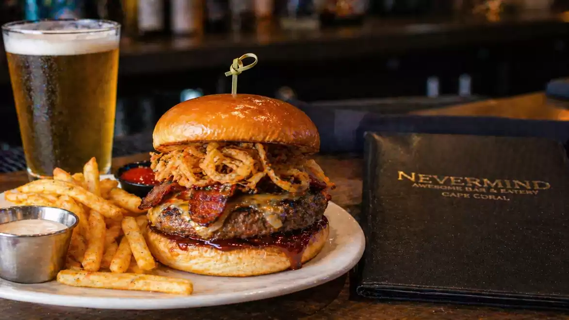 Juicy burger with onion rings, bacon, and fries on a plate next to a glass of beer in a restaurant.