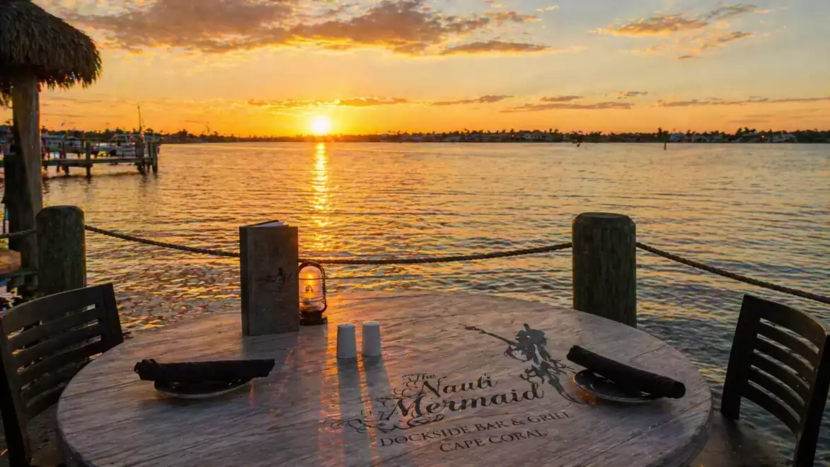 A round table with two chairs outdoors, overlooking calm waters with a sunset in the background. Wooden posts and a lantern adorn the scene.