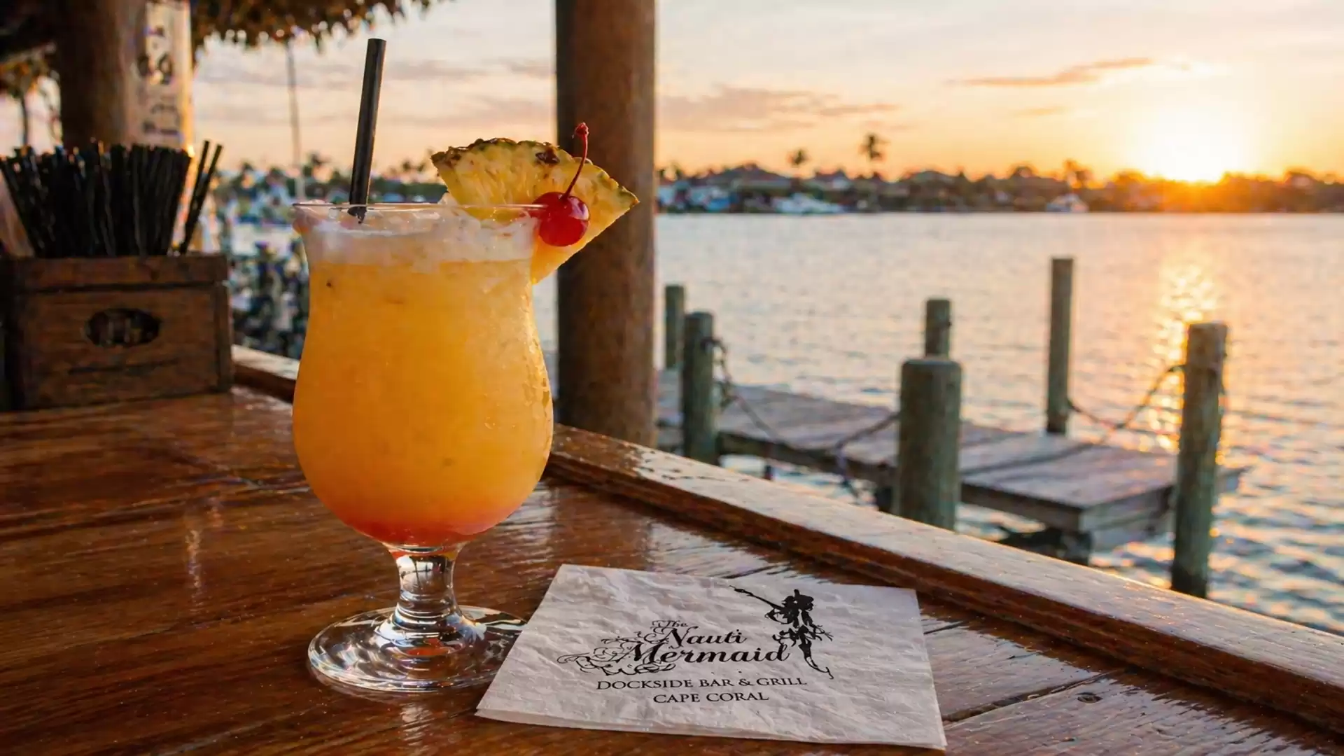 A tropical drink with pineapple and cherry on a wooden deck at sunset overlooking a waterfront.