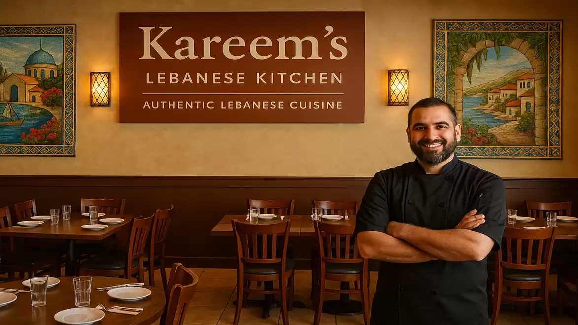 A smiling man stands with crossed arms in a cozy Lebanese restaurant named Kareem's Lebanese Kitchen. In the background, decorative artwork and set tables are visible.