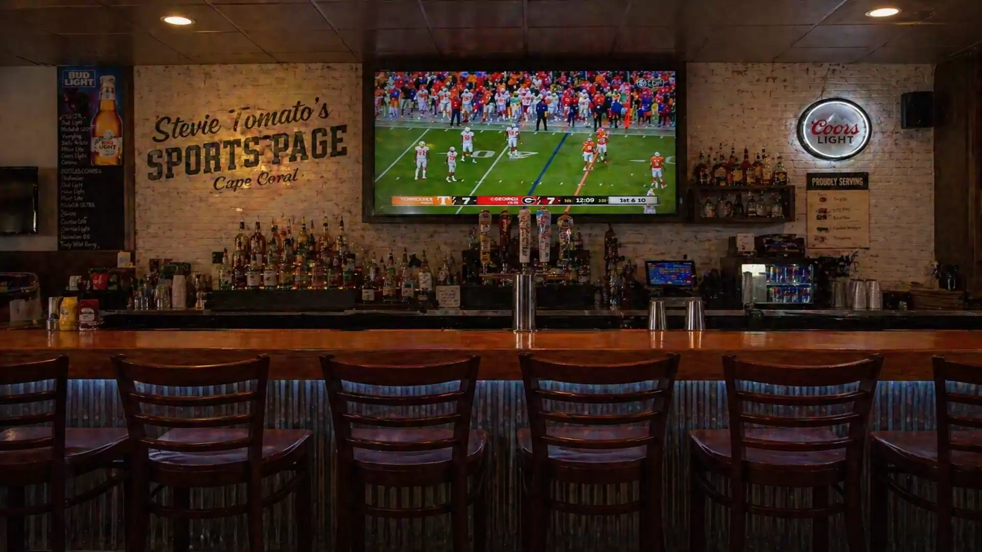 A bar with several high chairs in front of the counter. Behind it is a large flat-screen TV displaying a football game. A sign on the wall reads 'Stevie Tomato's Sportspage'. The bar is dimly lit, showcasing numerous bottles of spirits.