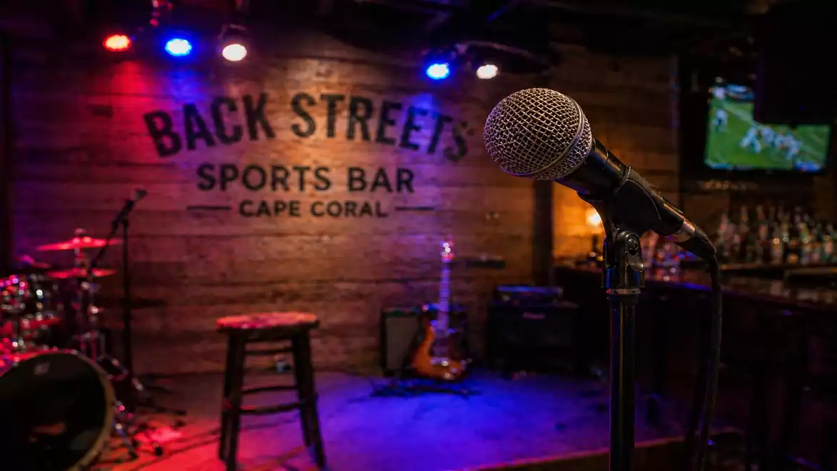 A cozy stage in a bar with a microphone in the foreground, colorful lights, and instruments in the background.