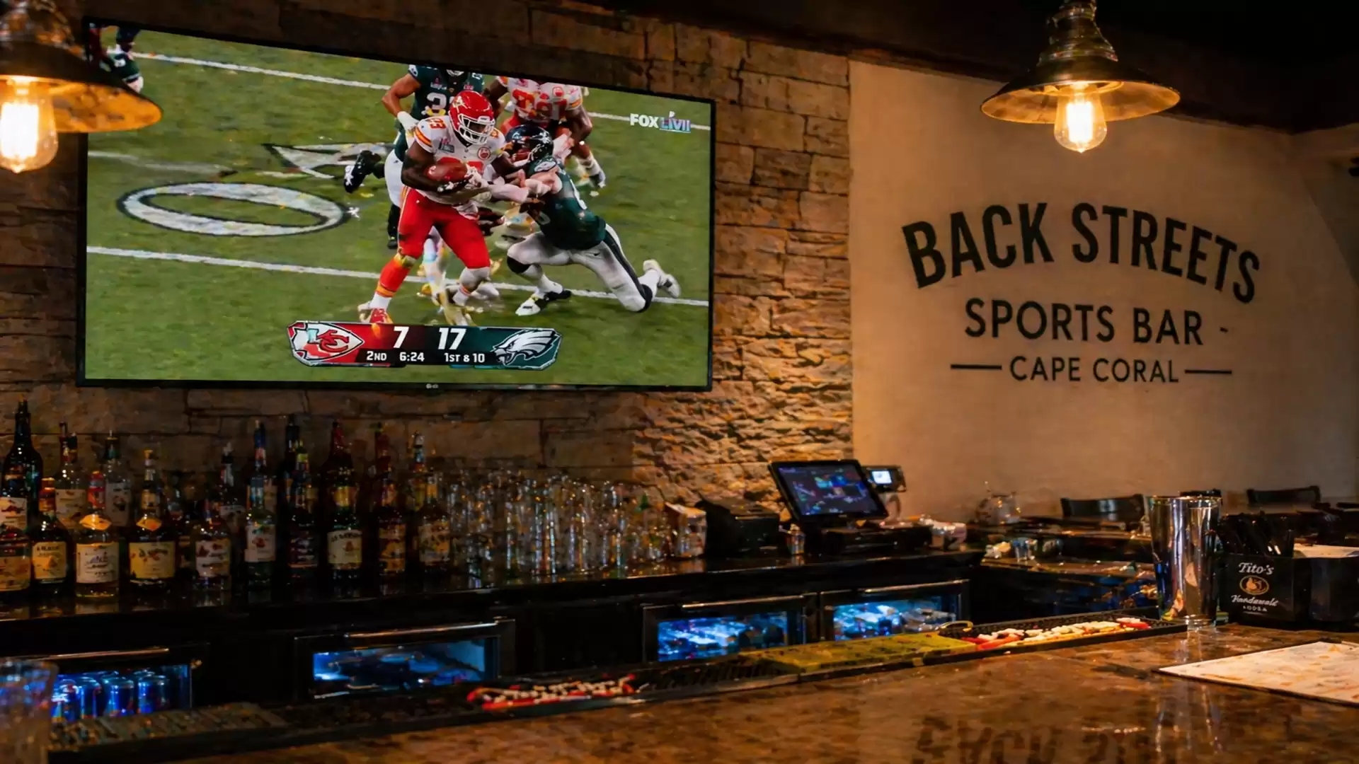 Image of a sports bar with a TV displaying an American football game. The bar with bottles and glasses is visible in the background.