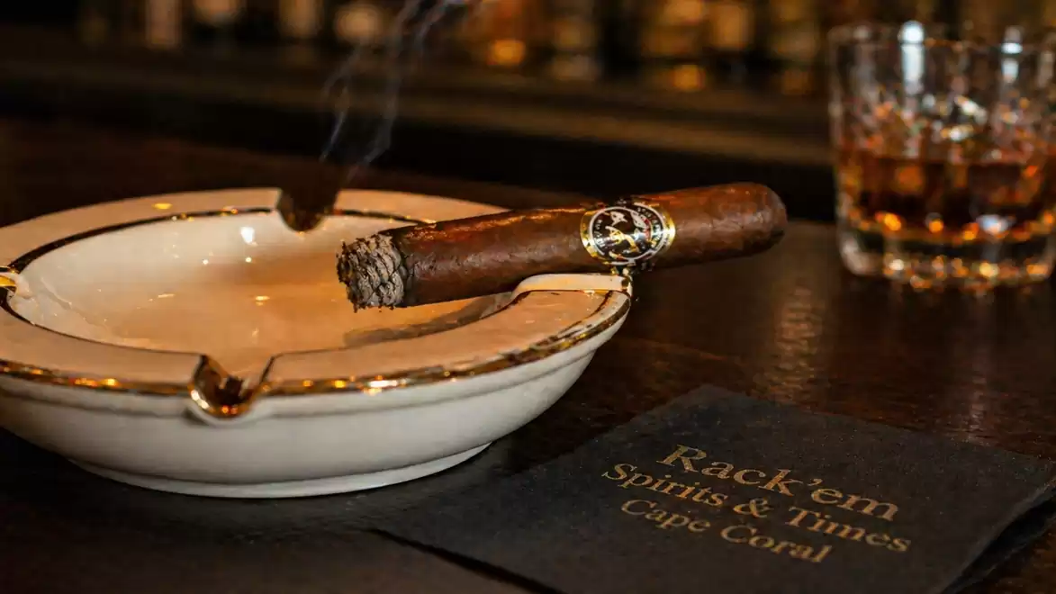 A lit cigar rests on an ashtray in a bar. In the background, a glass of whiskey is visible.