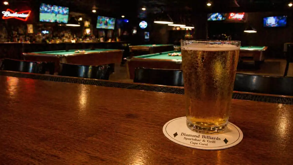 A glass of beer sits on a wooden counter in a bar with several pool tables in the background.