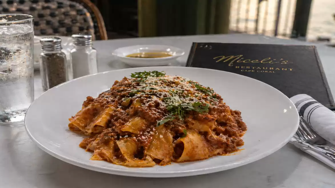A plate of fresh pasta in a meat sauce, garnished with Parmesan and parsley, sits on a set table with a menu in the background.