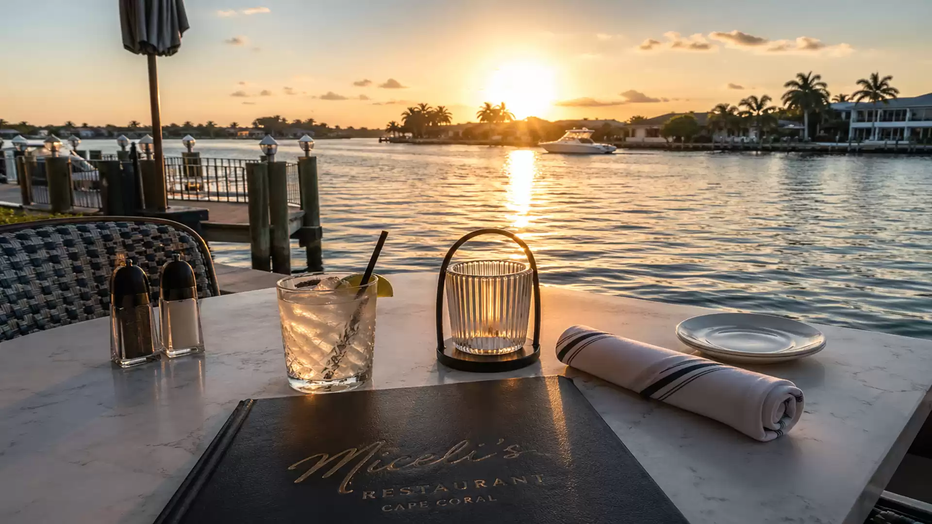 Sunset over a calm river with a set table in the foreground, featuring a drink, a candle, and cutlery.