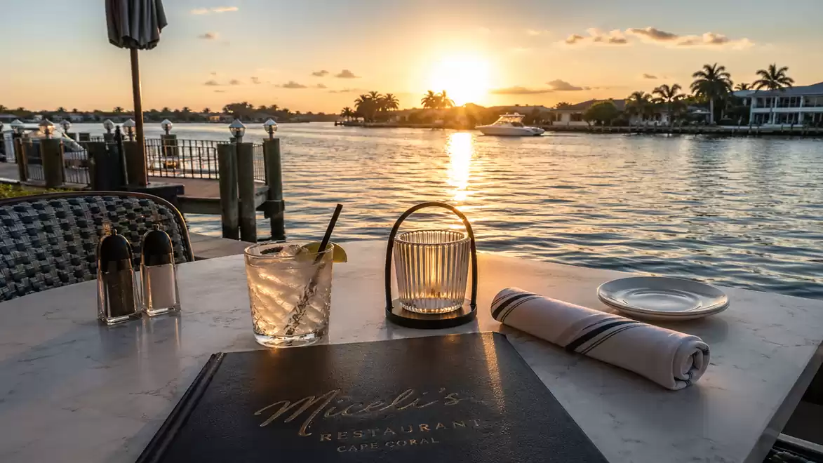 Sunset over a calm river with a set table in the foreground, featuring a drink, a candle, and cutlery.