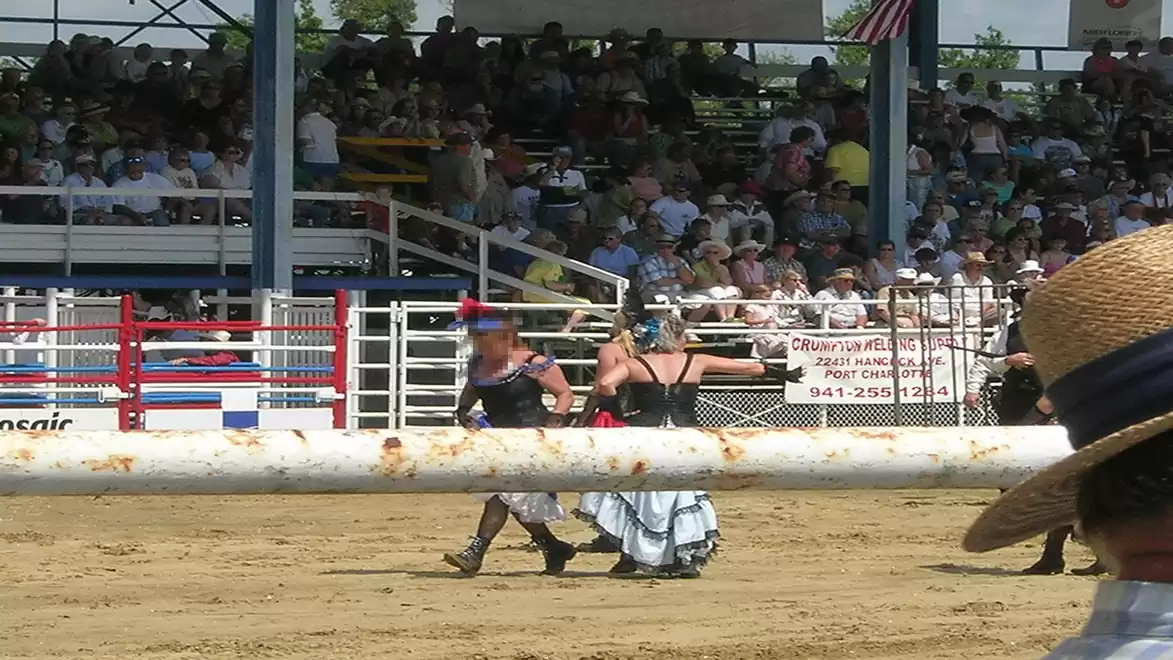 Two costumed participants at a rodeo event walk across the sandy area while spectators watch from the stands. A large straw hat is visible in the foreground.
