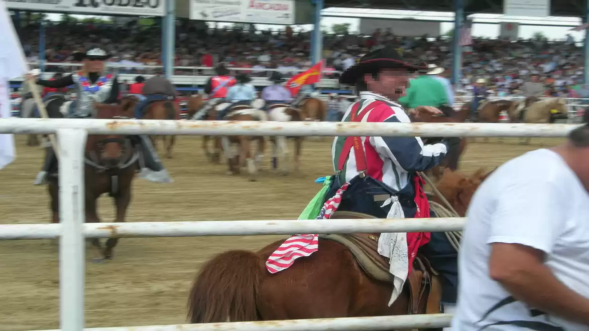 A cowboy in a rodeo, sitting on a horse surrounded by a wooden fence. In the background, there are other riders and a crowded stadium.