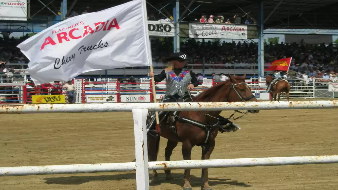 A rider on a horse holds a white flag with the text 'Arcadia Trucks' in a rodeo arena.