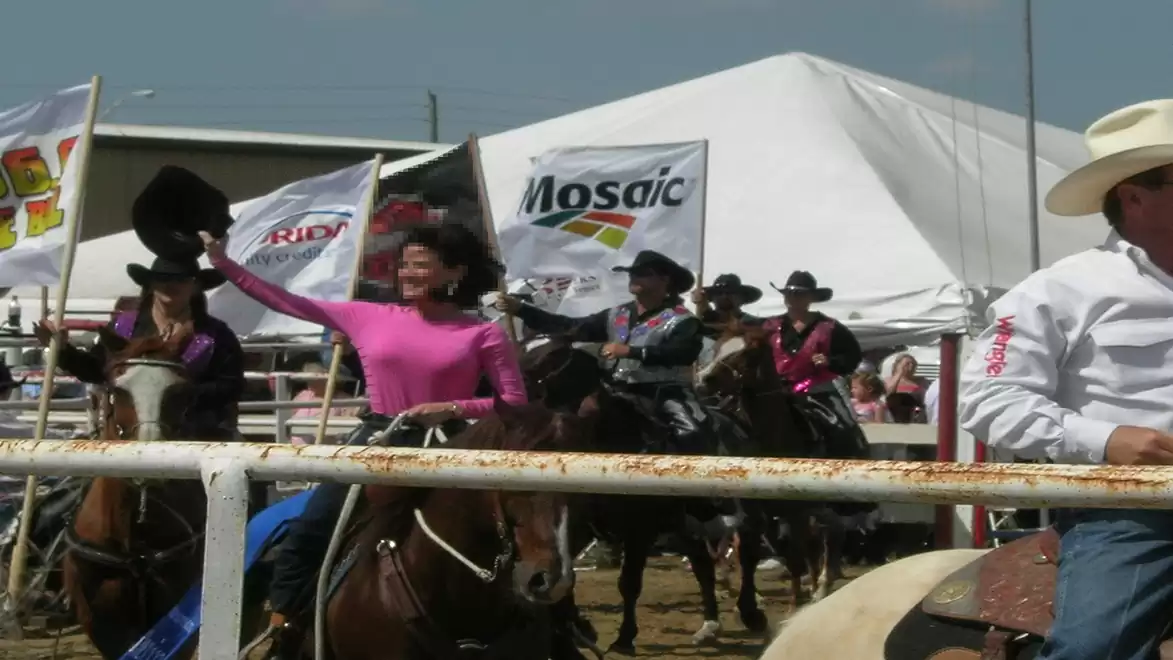 A group of people dressed as cowboys riding horses at a rodeo event. A woman in a pink shirt raises her cowboy hat. Tents and flags are visible in the background.