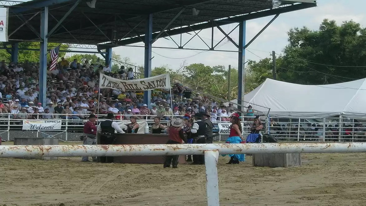 A rural event with spectators in bleachers and a busy area in the center marked by a banner. People are dressed in Western attire, and there are trees in the background.