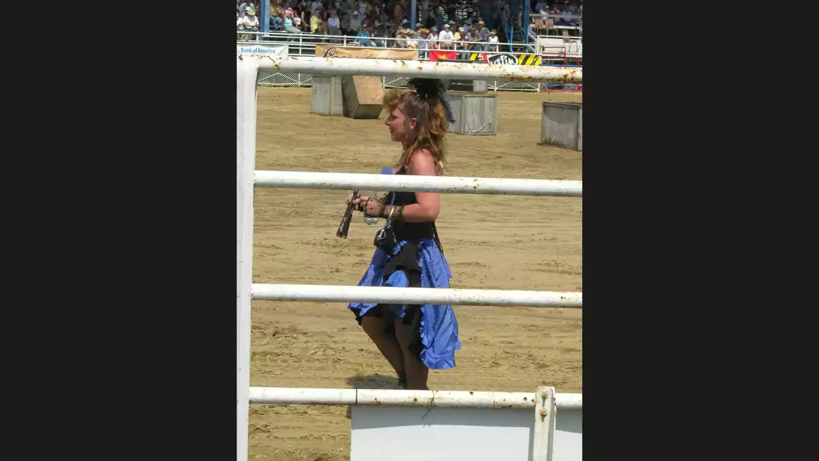 A woman in a blue dress walks behind a metal fence at an outdoor event. Spectators are visible in the background.