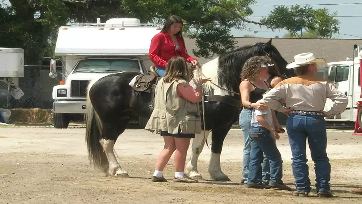 Woman sitting on a black horse, surrounded by several people outdoors, next to a parked RV.