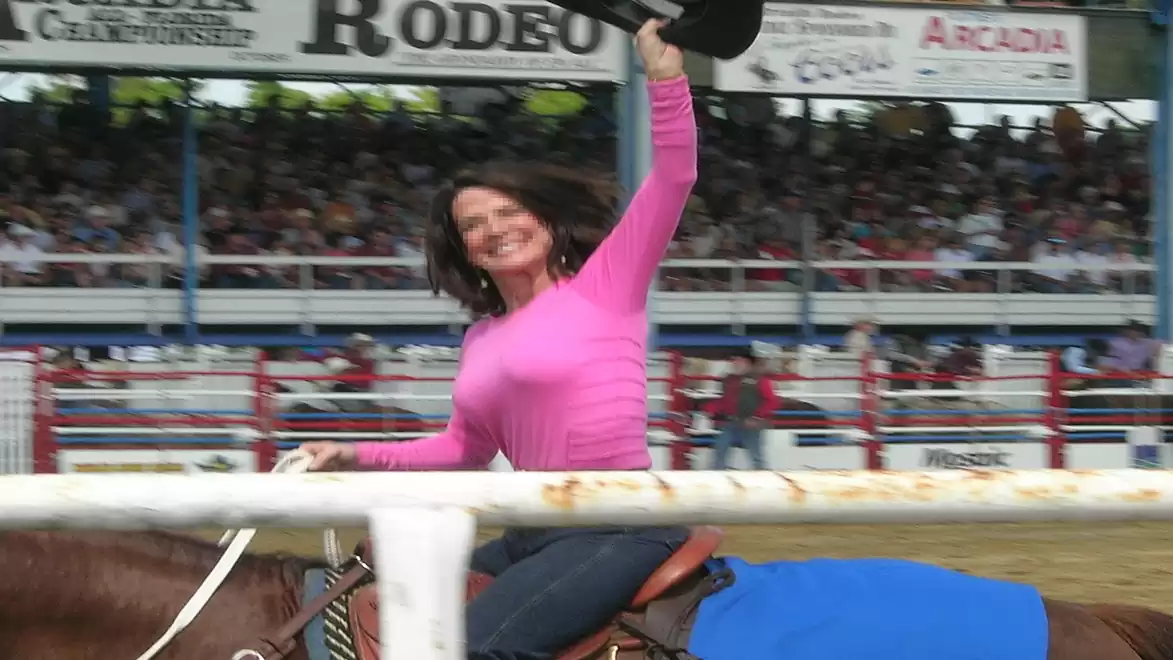 Woman in a pink sweater riding a horse at a rodeo event, waving a hat.