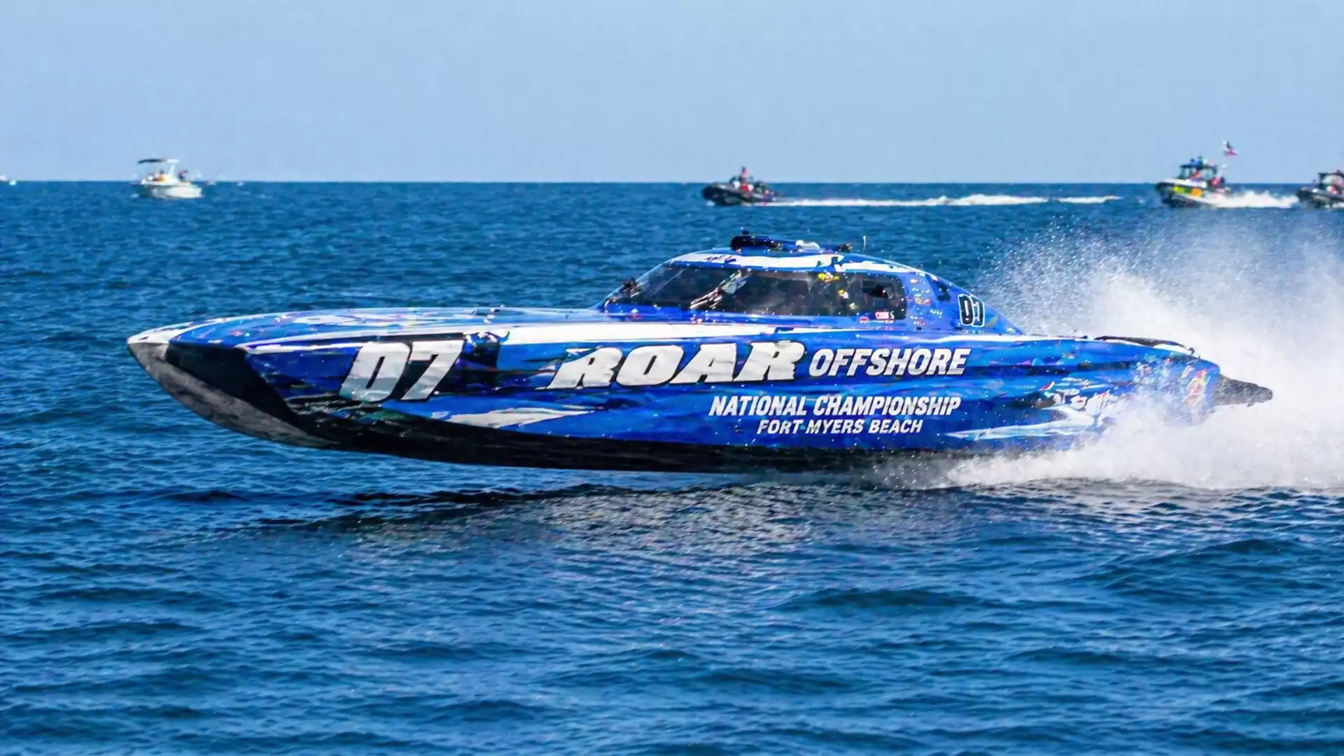 A blue offshore racing boat numbered 07 speeds across the sea, with other boats visible in the background.
