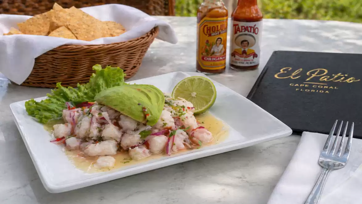 A deliciously presented ceviche with avocado slices and lime, served on a white plate next to hot sauce bottles and a basket with tortilla chips.