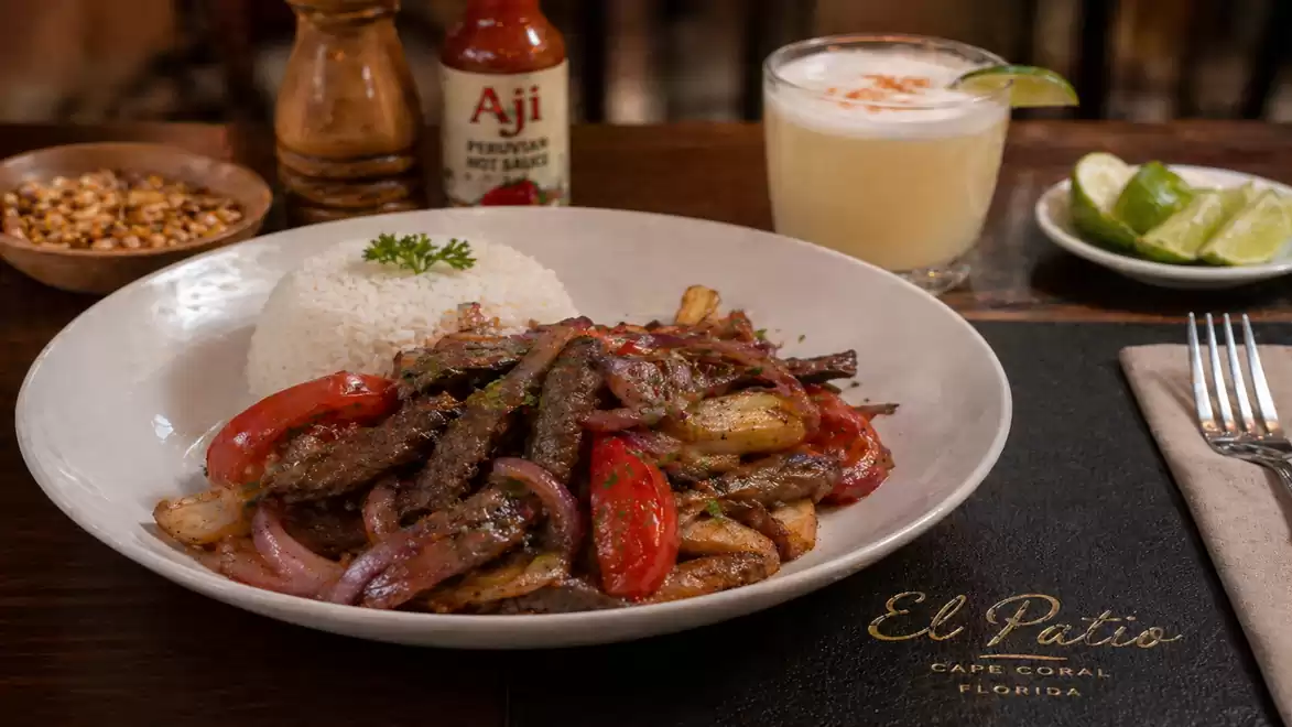 A traditional Peruvian dish featuring sautéed beef, tomatoes, onions, and potatoes, served with a portion of rice. In the background, there is a bottle of Aji sauce, a drink, and lime wedges.