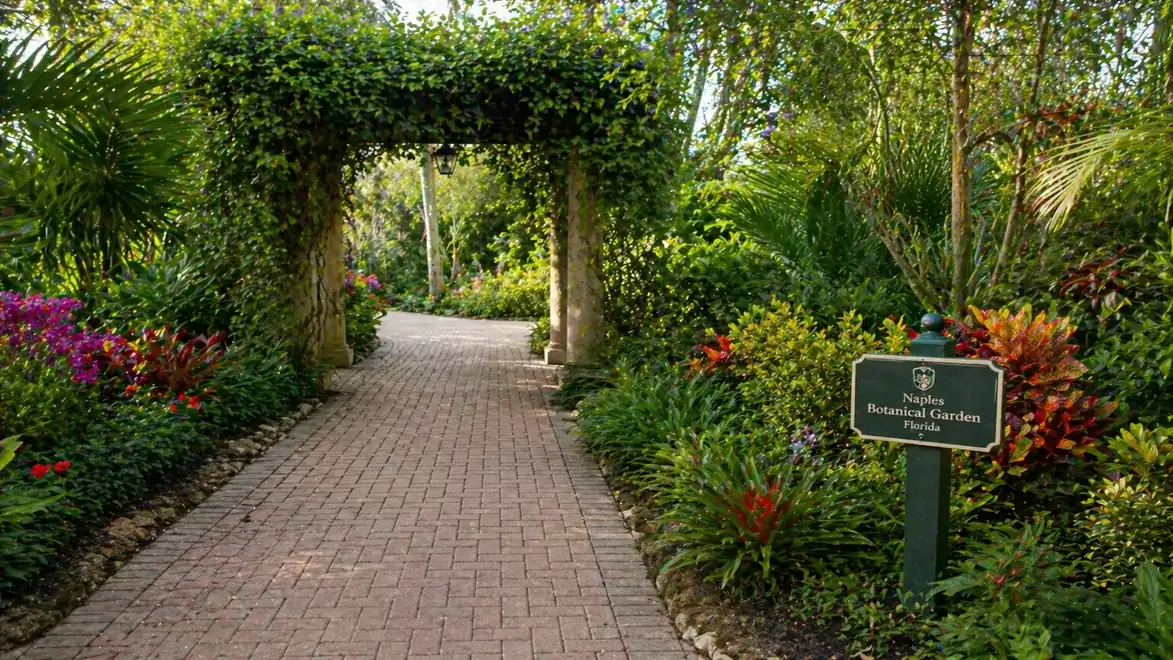 A paved path in a botanical garden leading through an ivy-covered archway, surrounded by lush plants and colorful flowers, with a sign on the right side.