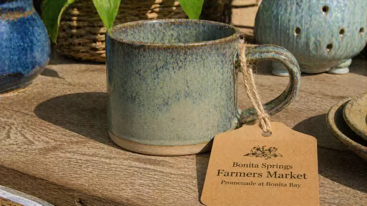 A handcrafted ceramic mug on a wooden table with a Bonita Springs Farmers Market tag, surrounded by other ceramic items.