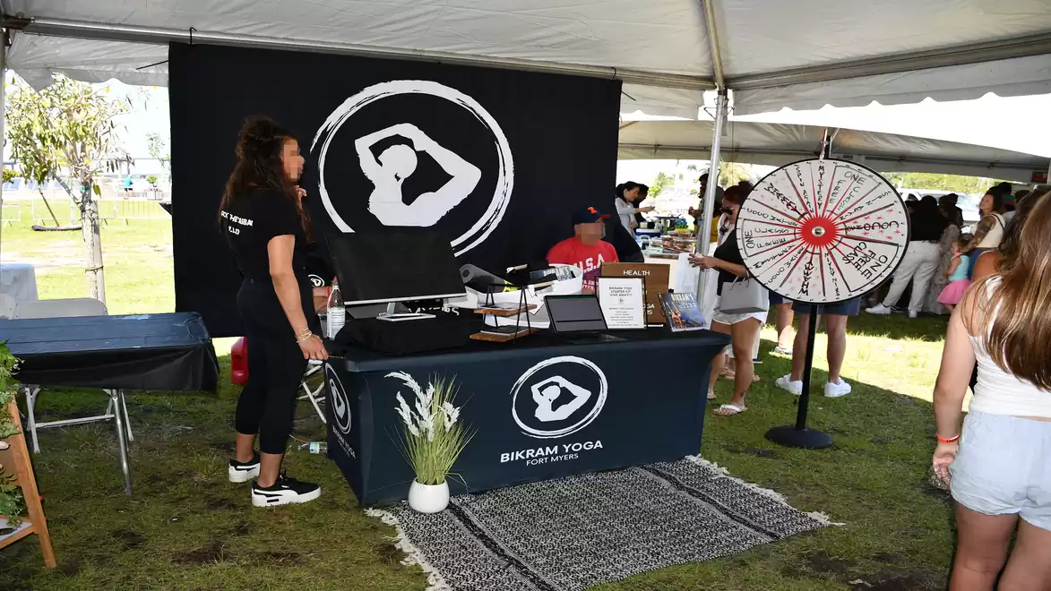 A booth with the Bikram Yoga logo on a table under a tent, surrounded by people. A spinning wheel is on the right in the image.