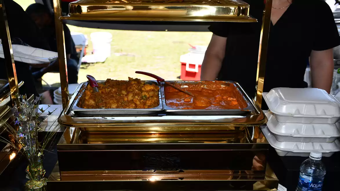 A buffet stand with a golden holder containing two warm dishes. On the left, there is a dish with potatoes and vegetables, and on the right, a curry dish. Nearby are some stacked disposable containers and a bottle of water.
