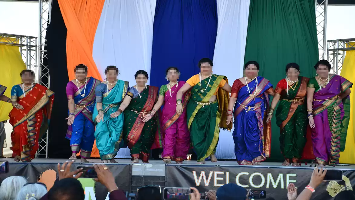 A group of people in traditional Indian clothing stands on a stage. Colorful fabric panels hang in the background.