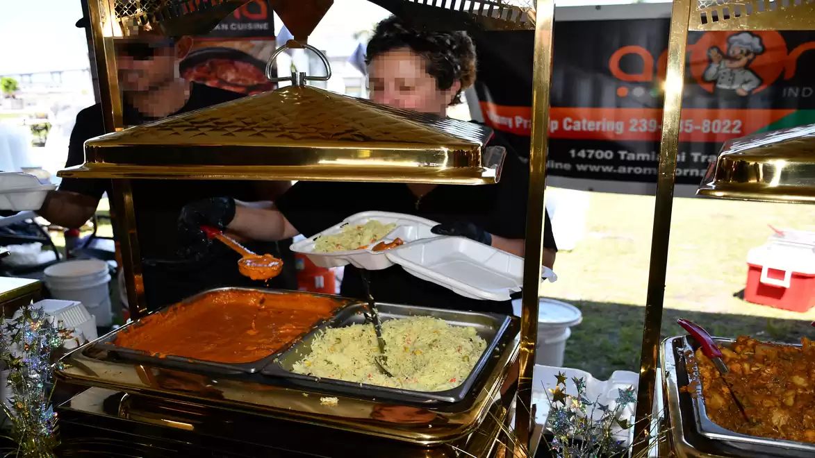 A catering booth with Indian food serving warm dishes at a buffet. A person is serving pasta and curry onto a plate.