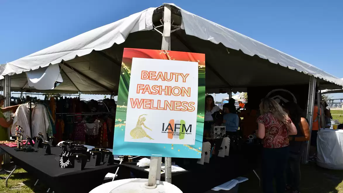 A large pavilion on a lawn with several stalls underneath. In the foreground is a sign reading 'Beauty Fashion Wellness' with the IAFM logo. Several people can be seen inside the pavilion while products are displayed on the tables.