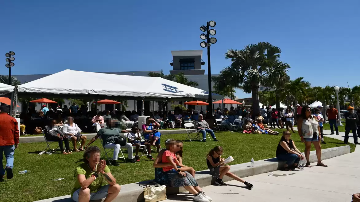 People relaxing in sunny weather in front of a large white tent with palm trees in the background.