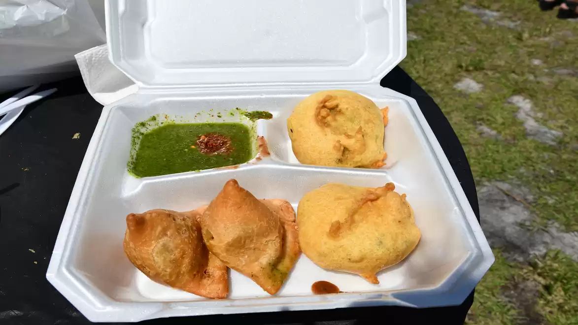 A white styrofoam container with two samosas and two fried balls, along with a green sauce, on a dark table outdoors.