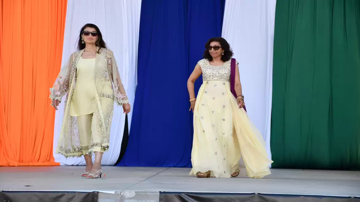 Two women in elegant clothing walk across a stage with a colorful background of orange, white, blue, and green.