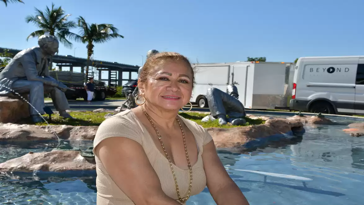 A woman is sitting and smiling by an outdoor fountain; in the background, palm trees, a delivery van, and a group of sculptures are visible.