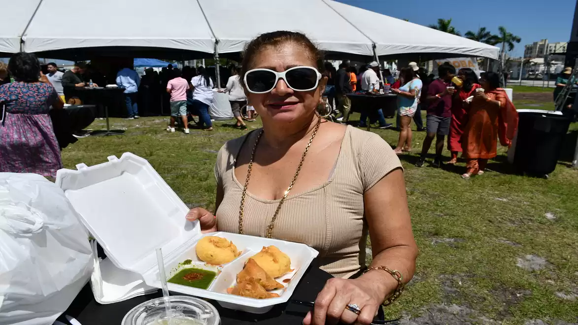Woman wearing sunglasses holding a food box with fried snacks in front of a white event tent at an outdoor gathering.