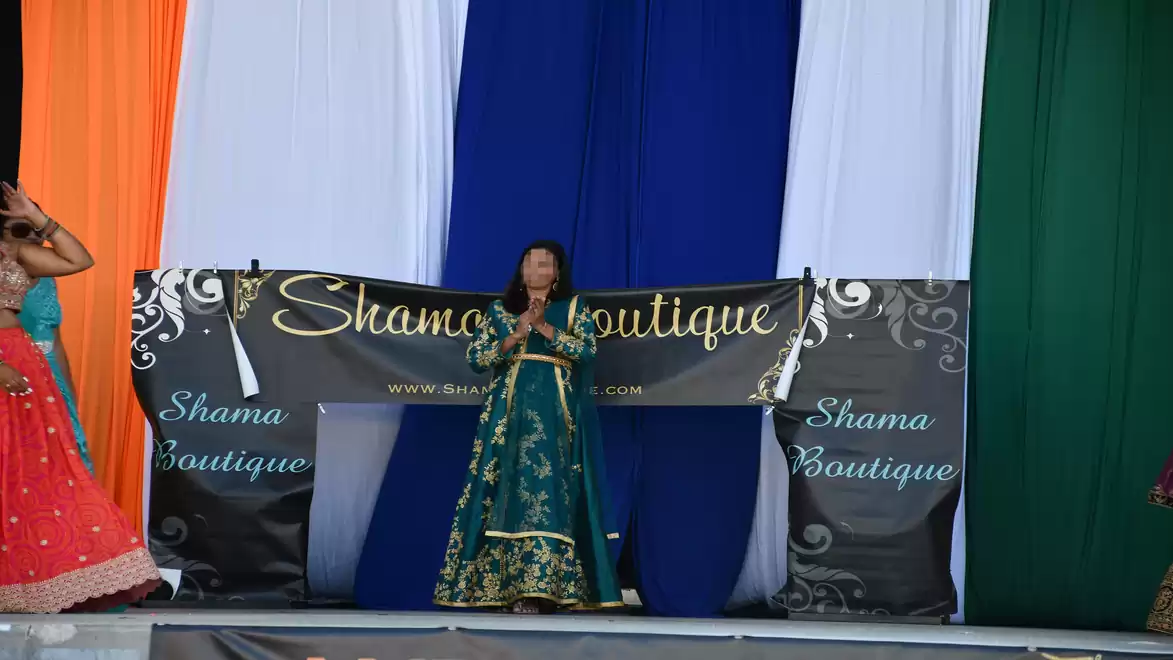 A woman in traditional clothing stands on a stage in front of a banner reading 'Shama Boutique'. Colorful curtains form the background.