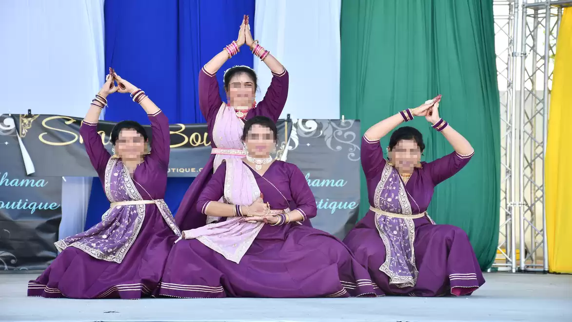 Four women in traditional purple dance costumes perform a classical choreography on a stage.