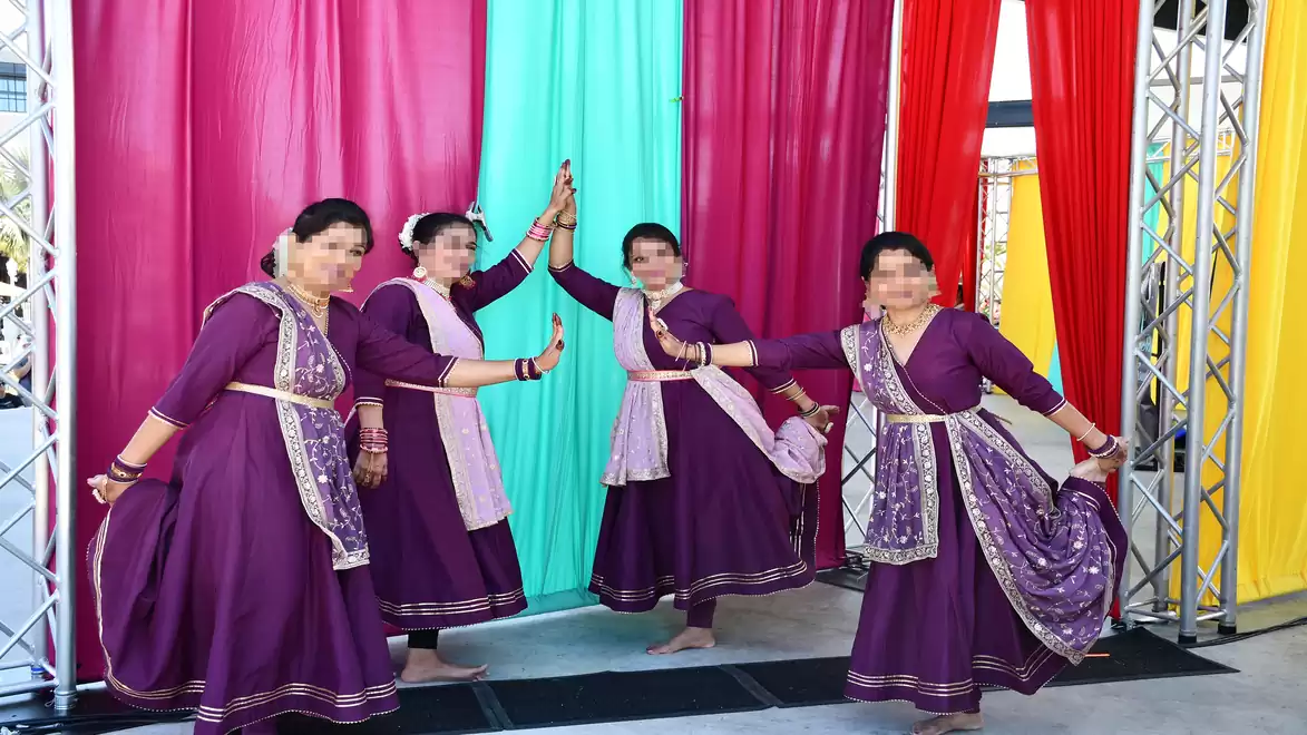 Four women in traditional purple dresses dancing and posing in front of colorful curtains.