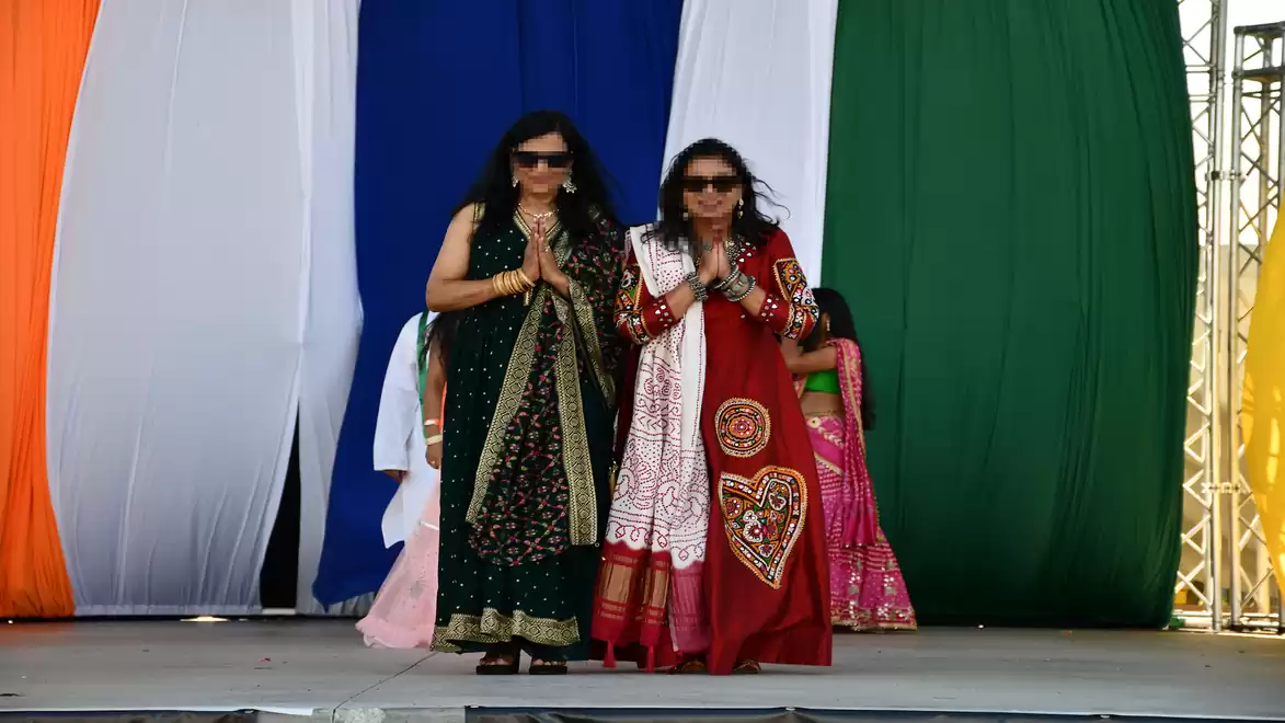 Two women in traditional Indian dresses walk onto a stage with a colorful curtain in the background.