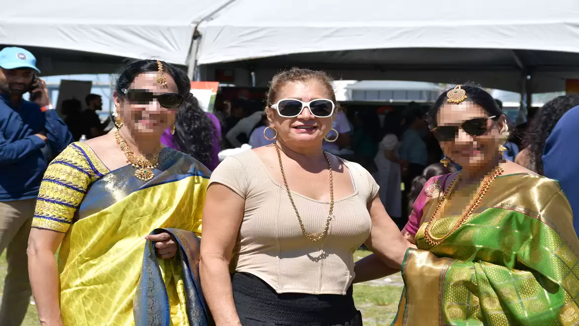 Three women smiling in traditional outfits at an outdoor event, under a large white tent.