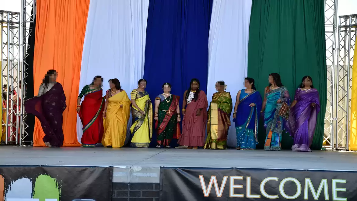 Group of women in traditional Indian saris standing on a stage with a backdrop in the colors orange, white, blue, and green.