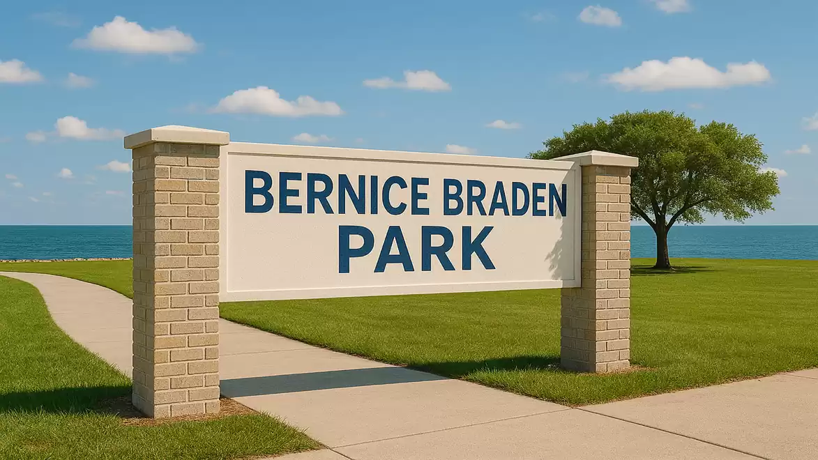 A sign reading 'Bernice Braden Park' is set on a well-maintained lawn with a tree and the sea in the background.