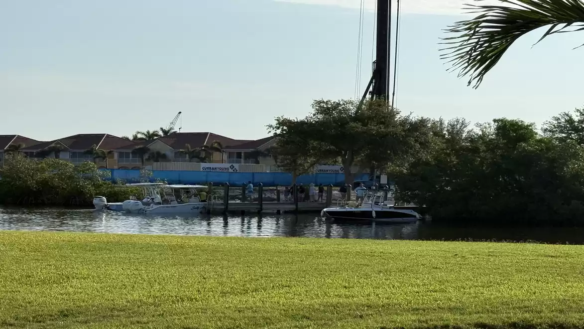 A calm river with boats docked, flanked by houses and palm trees in the background on a sunny day.