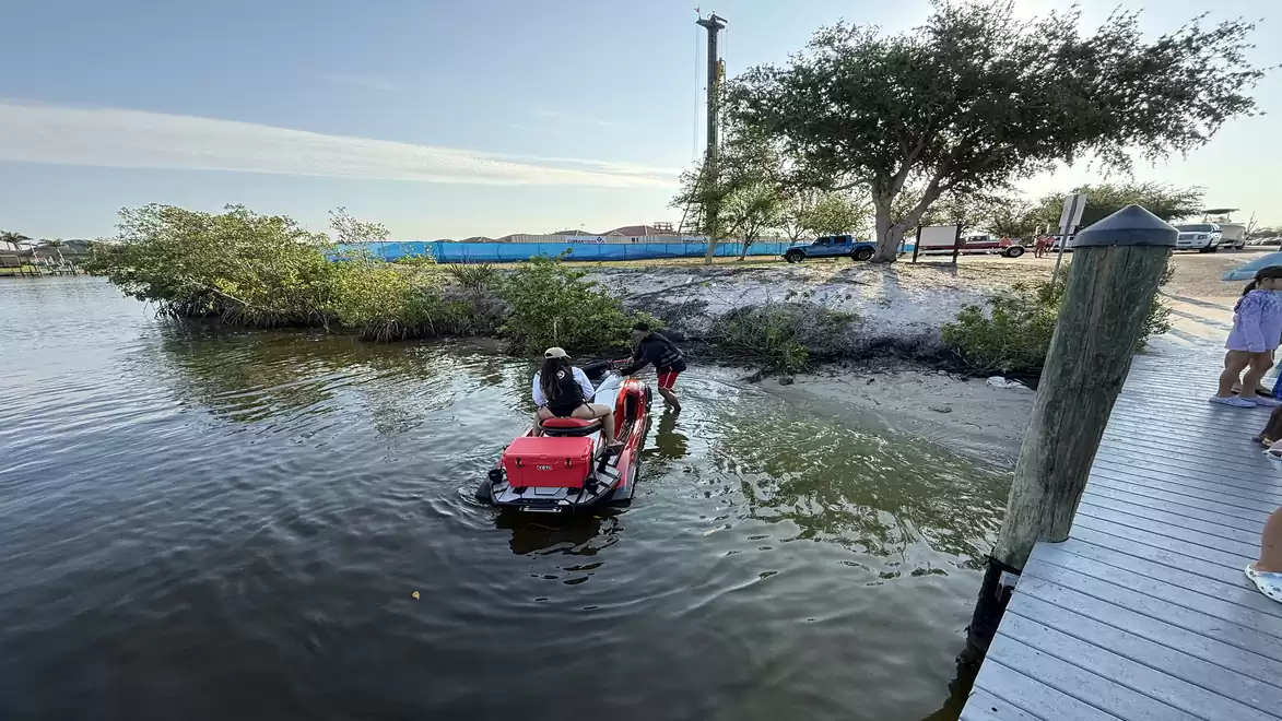 A jet ski rides on calm water near a dock with trees and people in the background.